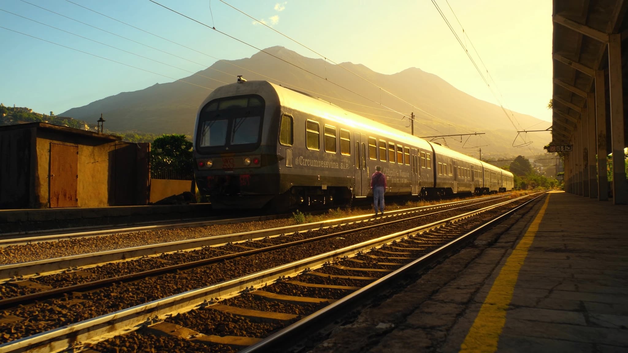 A Circumvesuviana commuter train at a station platform with Mount Vesuvius behind, golden hour