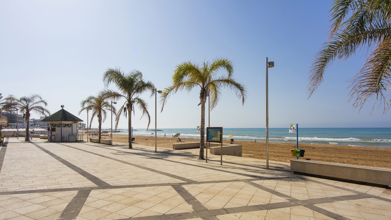 The paved seafront promenade at Marina di Ragusa, palms along the walkway and the sandy beach and open sea to the right